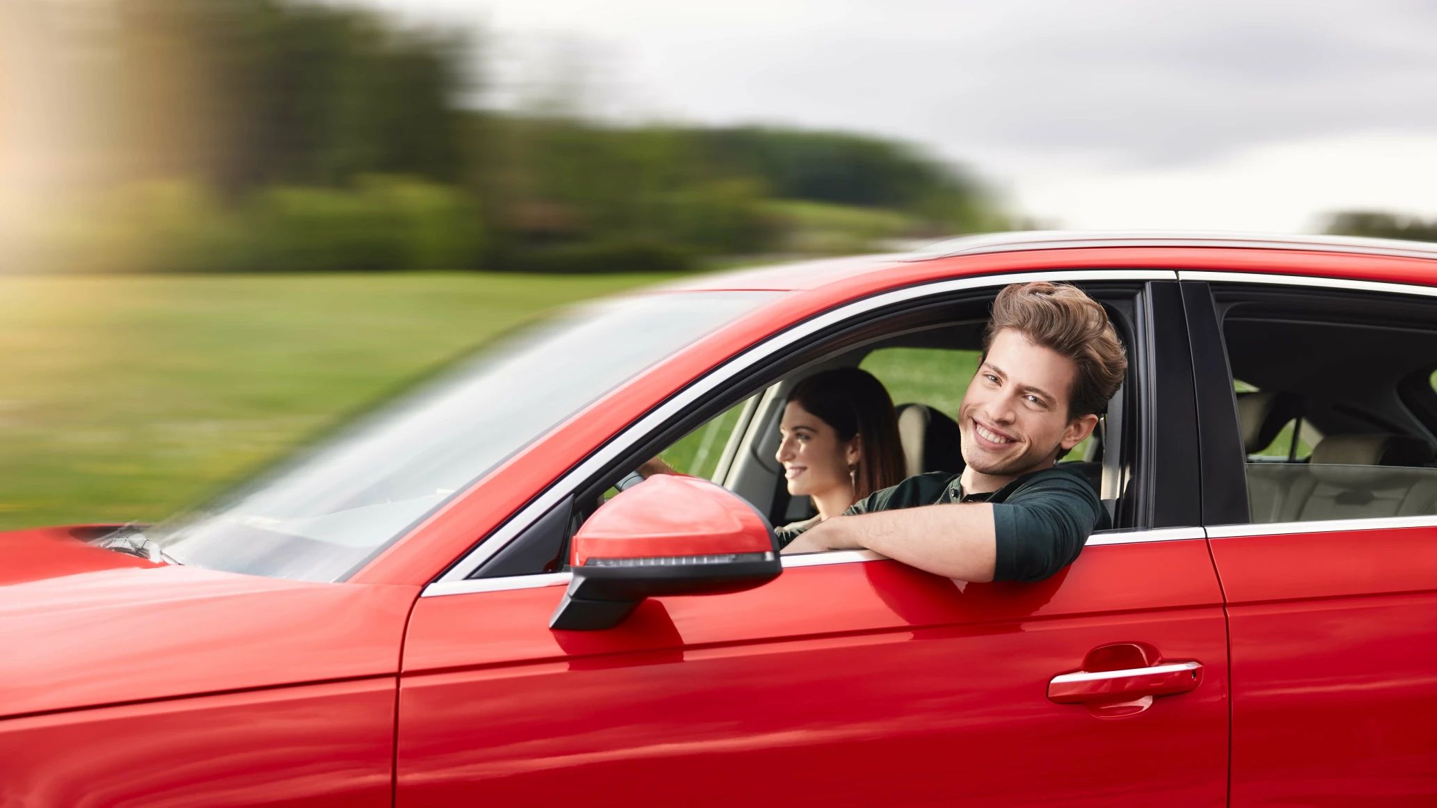 Ein junger Mann und eine Frau fahren in einem roten Auto durch die Natur und lächeln aus dem Auto.